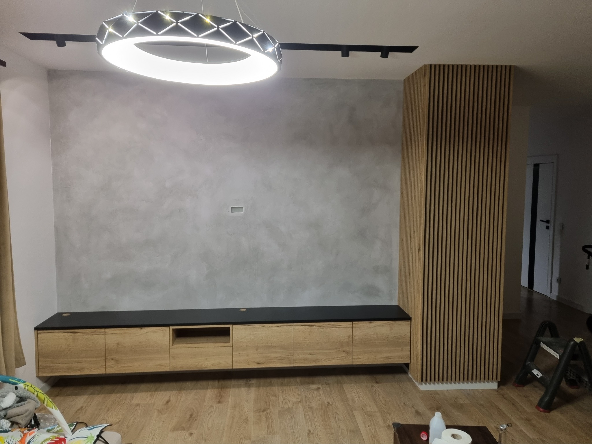 Nowoczesny salon z drewnianą zabudową TV i lamelami ściennymi. Minimalistyczny design, oświetlenie punktowe i surowa ściana tworzą unikalny klimat wnętrza.