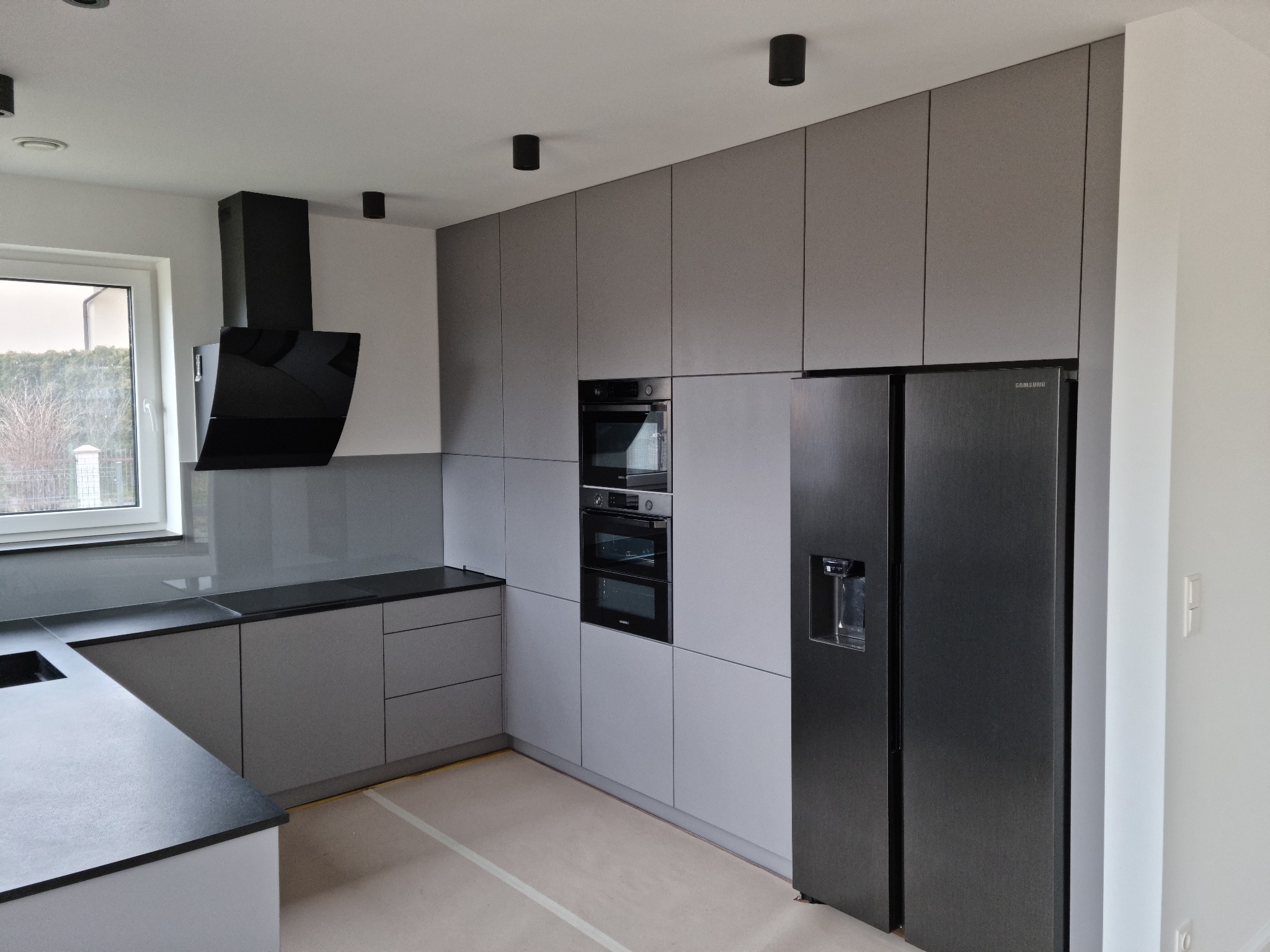 Nowoczesna kuchnia w odcieniach szarości z czarnymi akcentami: okap, piekarnik i lodówka side-by-side. Minimalistyczny design, gładkie fronty szafek, punktowe oświetlenie.