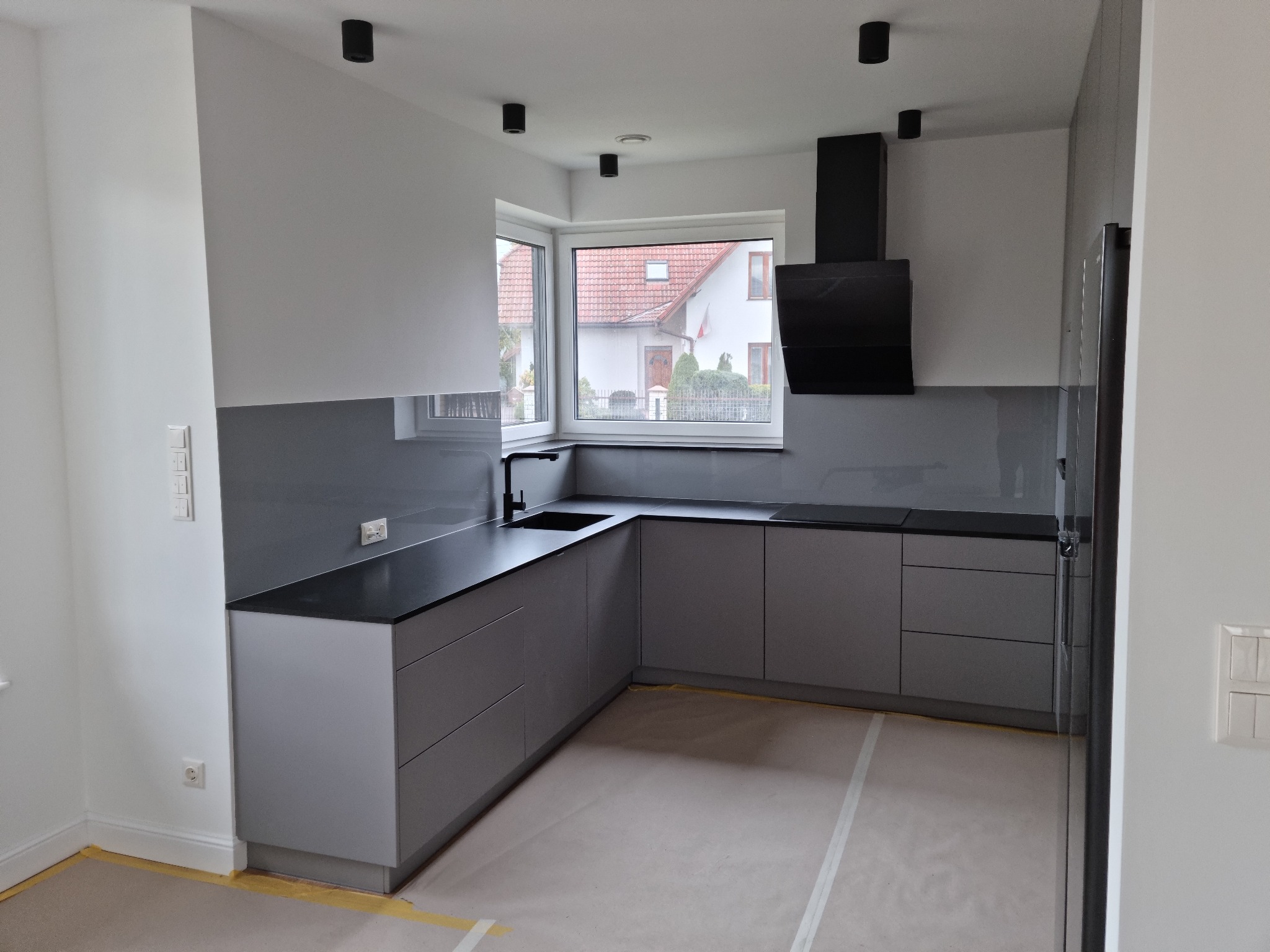 Nowoczesna, szara kuchnia z czarnym blatem i okapem, widok na dom przez okno. Podłoga zabezpieczona folią malarską, minimalistyczne oświetlenie sufitowe.