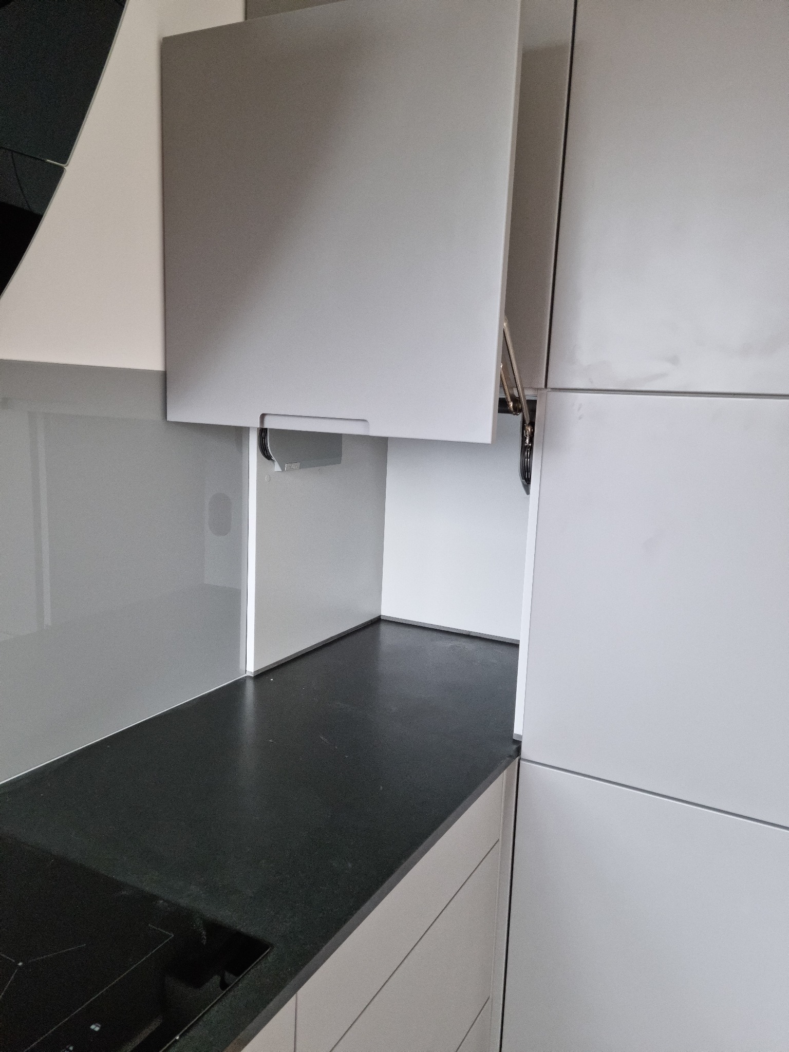 Nowoczesna kuchnia z szarymi szafkami i czarnym blatem. Szafka górna otwarta, ukazująca mechanizm podnoszenia. Minimalistyczny design, gładkie fronty.