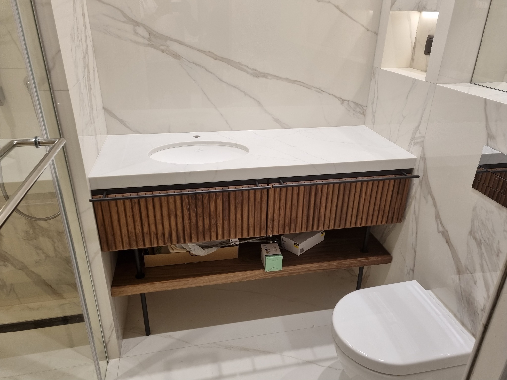 Nowoczesna łazienka z umywalką nablatową na szafce z lamelami i blatem z białego marmuru. W tle ściana wyłożona płytami imitującymi marmur.