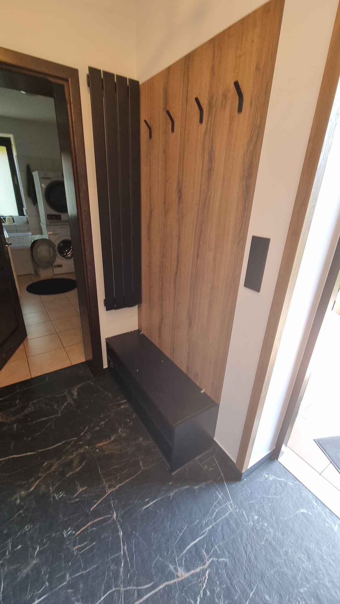 Nowoczesny przedpokój z drewnianą zabudową, czarnymi haczykami i grzejnikiem panelowym. Ciemna ławka i marmurowe płytki podłogowe. Widok na pralkę w tle.