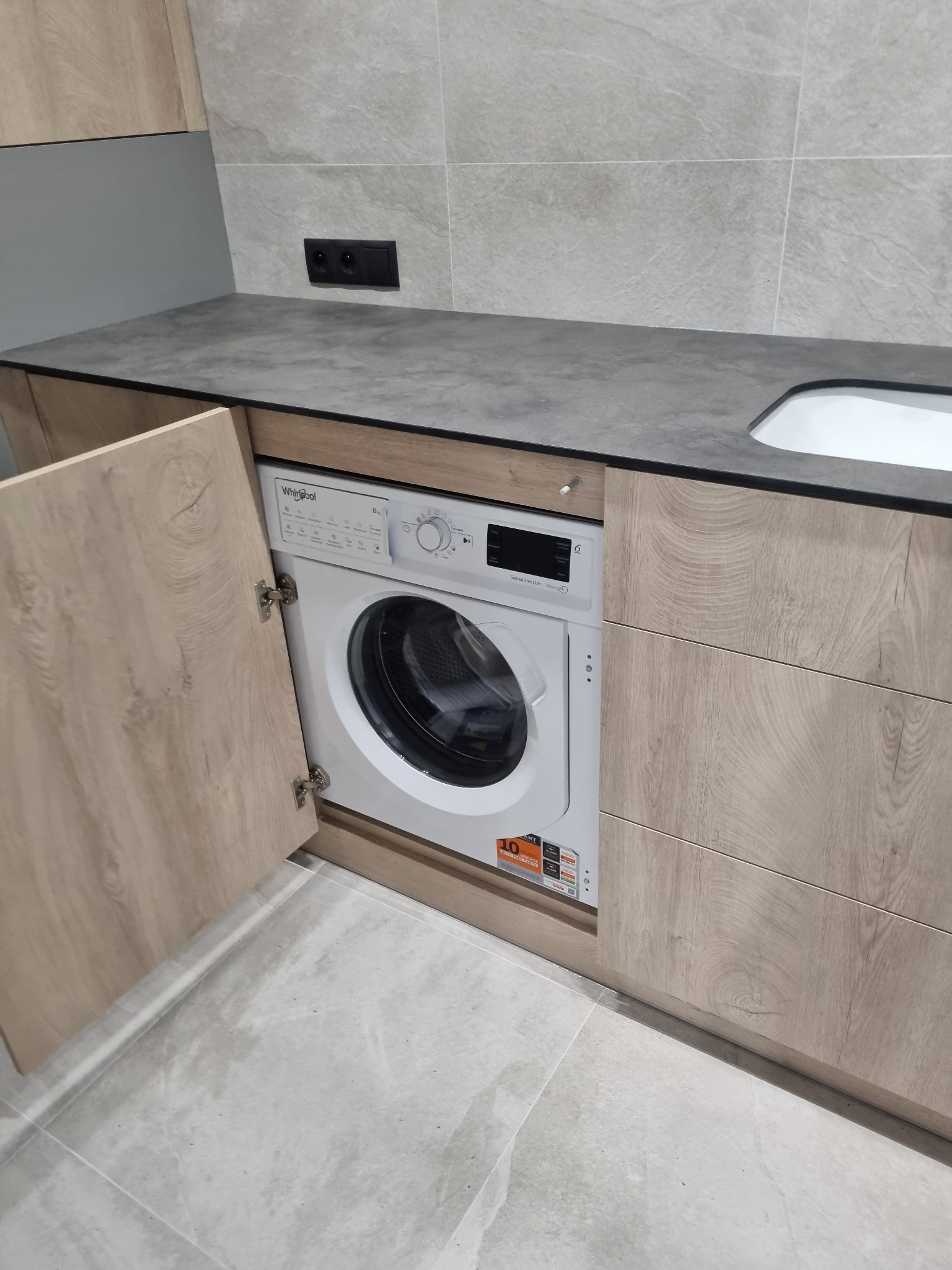 Biała pralka Whirlpool w zabudowie meblowej z frontem imitującym jasne drewno, szary blat i płytki. Otwarta szafka z lewej strony. Nowoczesny design.