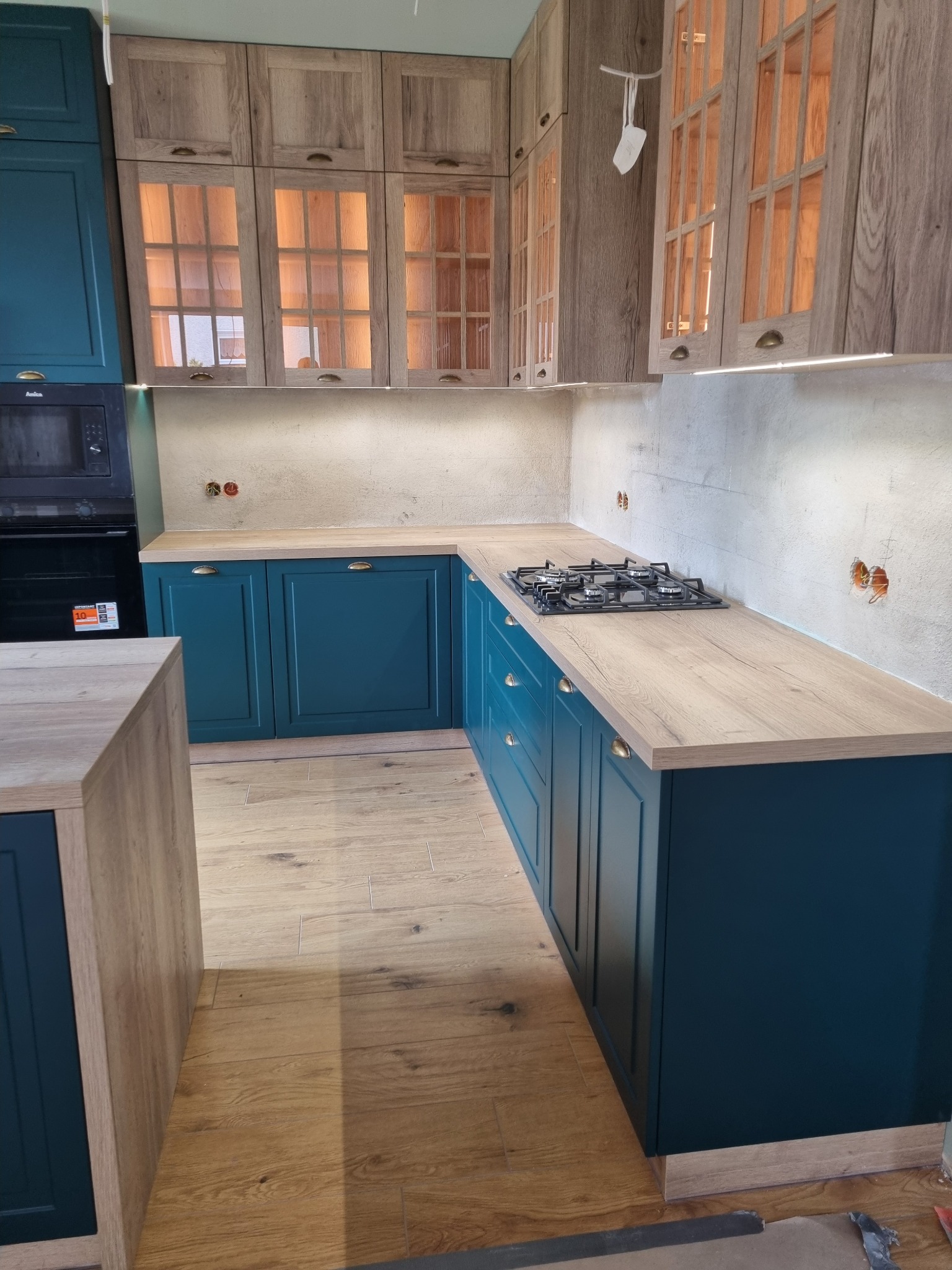 Nowoczesna kuchnia w odcieniach drewna i turkusu. Górne szafki z przeszkleniami, drewniany blat, płyta gazowa. Widoczne niedokończone wykończenia ścian.