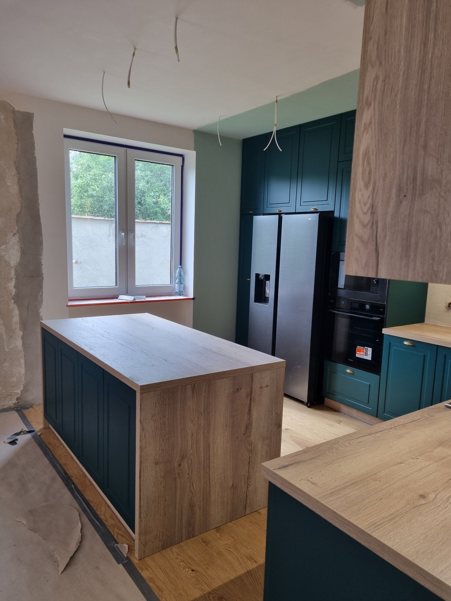 Wnętrze kuchni w trakcie wykańczania: wyspa z blatem drewnopodobnym i ciemnozielonymi frontami, pasujące szafki, sprzęt AGD. Widoczne niedokończone ściany i wystające kable.