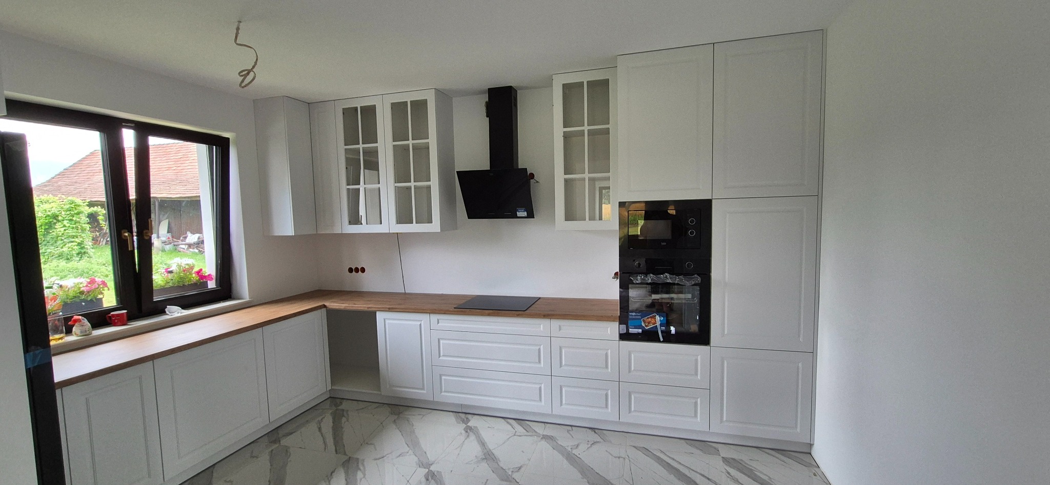 Jasna kuchnia z białymi szafkami i drewnianym blatem, okno z widokiem na zieleń. Nowoczesny design z czarnym okapem i marmurową podłogą.