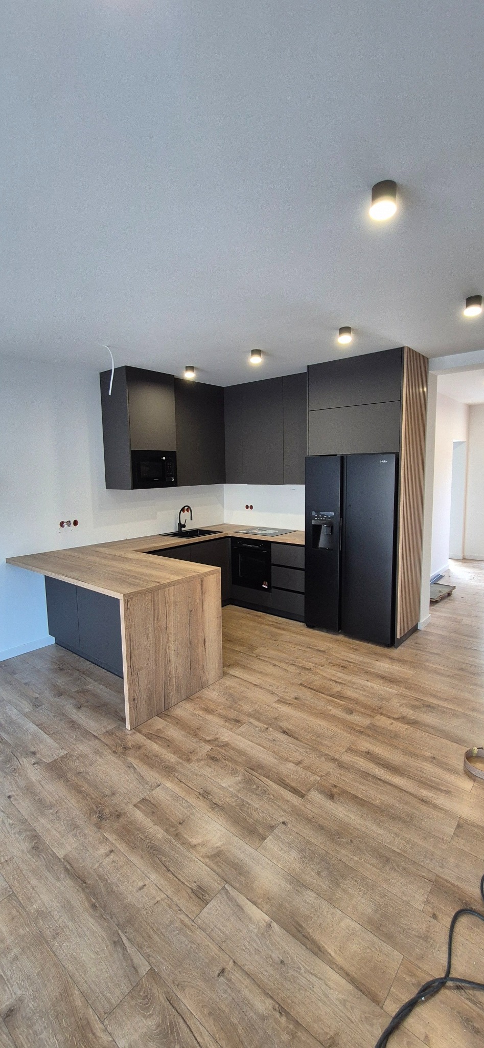 Nowoczesna, minimalistyczna kuchnia z czarnymi szafkami i drewnianym blatem. Widoczne oświetlenie punktowe i drewniane panele dekoracyjne obok lodówki.