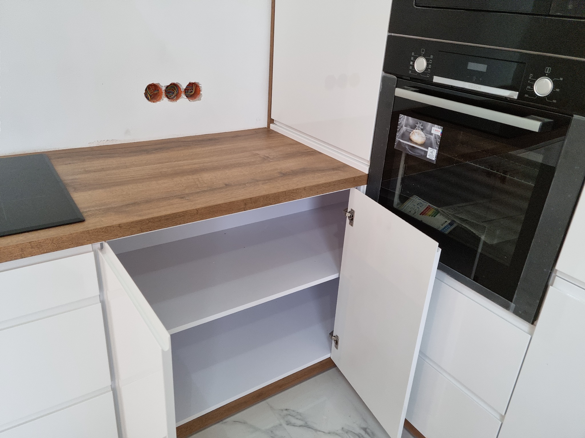 Fragment kuchni z otwartą szafką z białymi frontami i blatem imitującym drewno, obok zabudowana piekarnik. Widoczne niedokończone otwory na gniazdka elektryczne w ścianie.