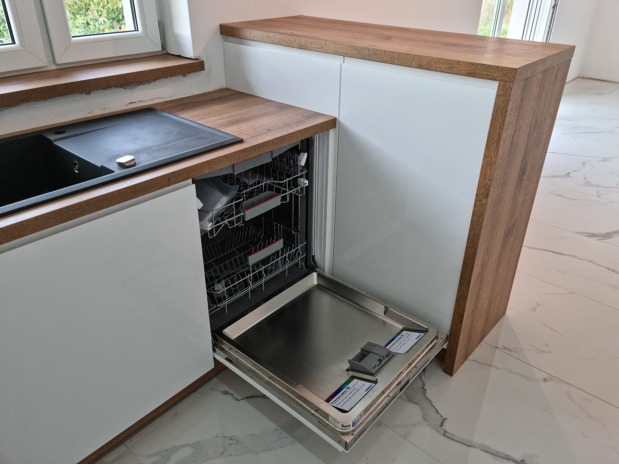 Nowoczesna kuchnia z białymi szafkami i drewnianym blatem, widoczna otwarta zmywarka z naklejkami producenta na drzwiczkach, marmurowa podłoga.