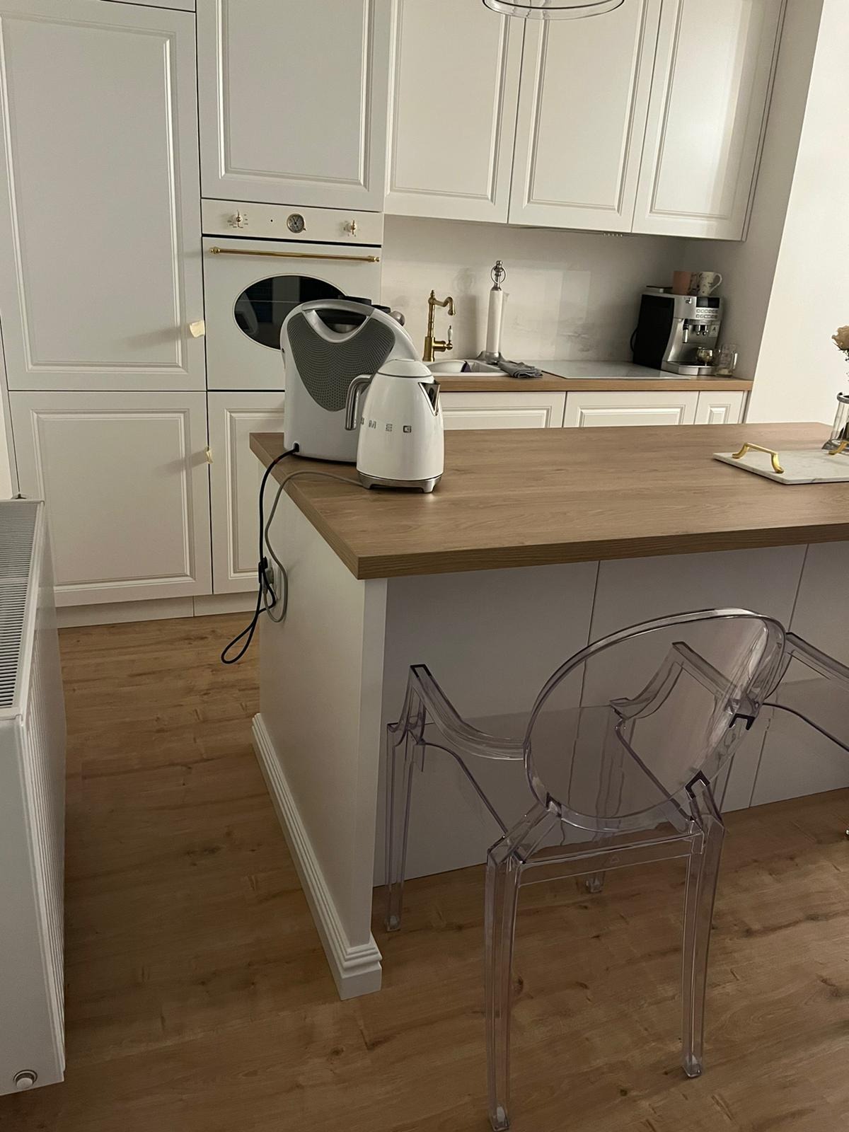 Biała kuchnia z drewnianym blatem wyspy, na której stoją biały czajnik elektryczny i toster marki Smeg, w tle ekspres do kawy i bateria kuchenna w kolorze złotym, z przodu przezroczyste krzesło...