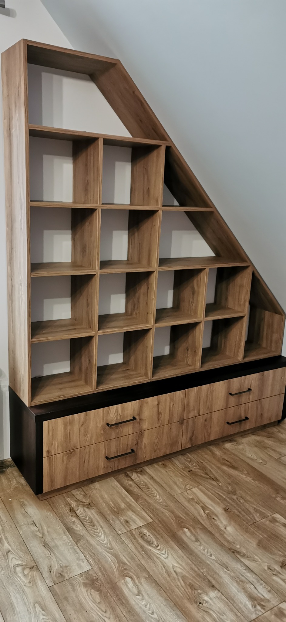 Drewniany regał z szufladami wkomponowany pod skos dachu, z widoczną strukturą drewna i czarnymi uchwytami szuflad, na podłodze z paneli.