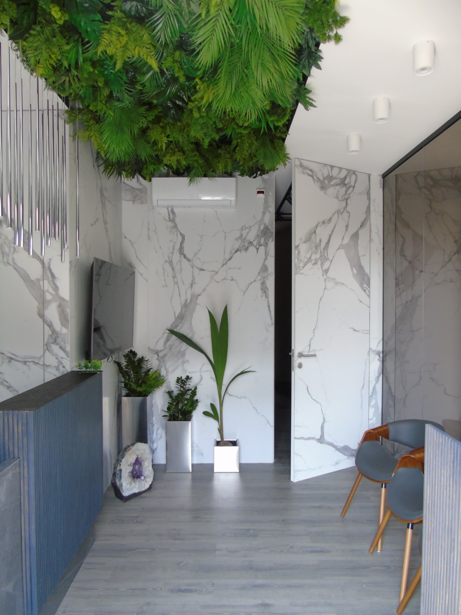 Nowoczesne wnętrze biura z marmurowymi ścianami, sztuczną roślinnością na suficie i szarymi panelami podłogowymi, uzupełnione minimalistycznymi meblami i dekoracjami.