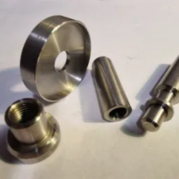 Cztery metalowe elementy o różnych kształtach, prawdopodobnie wykonane na tokarce lub obrabiarce CNC, leżące na jasnym tle. Jeden element to pierścień z otworem, drugi to tuleja, trzeci to element...