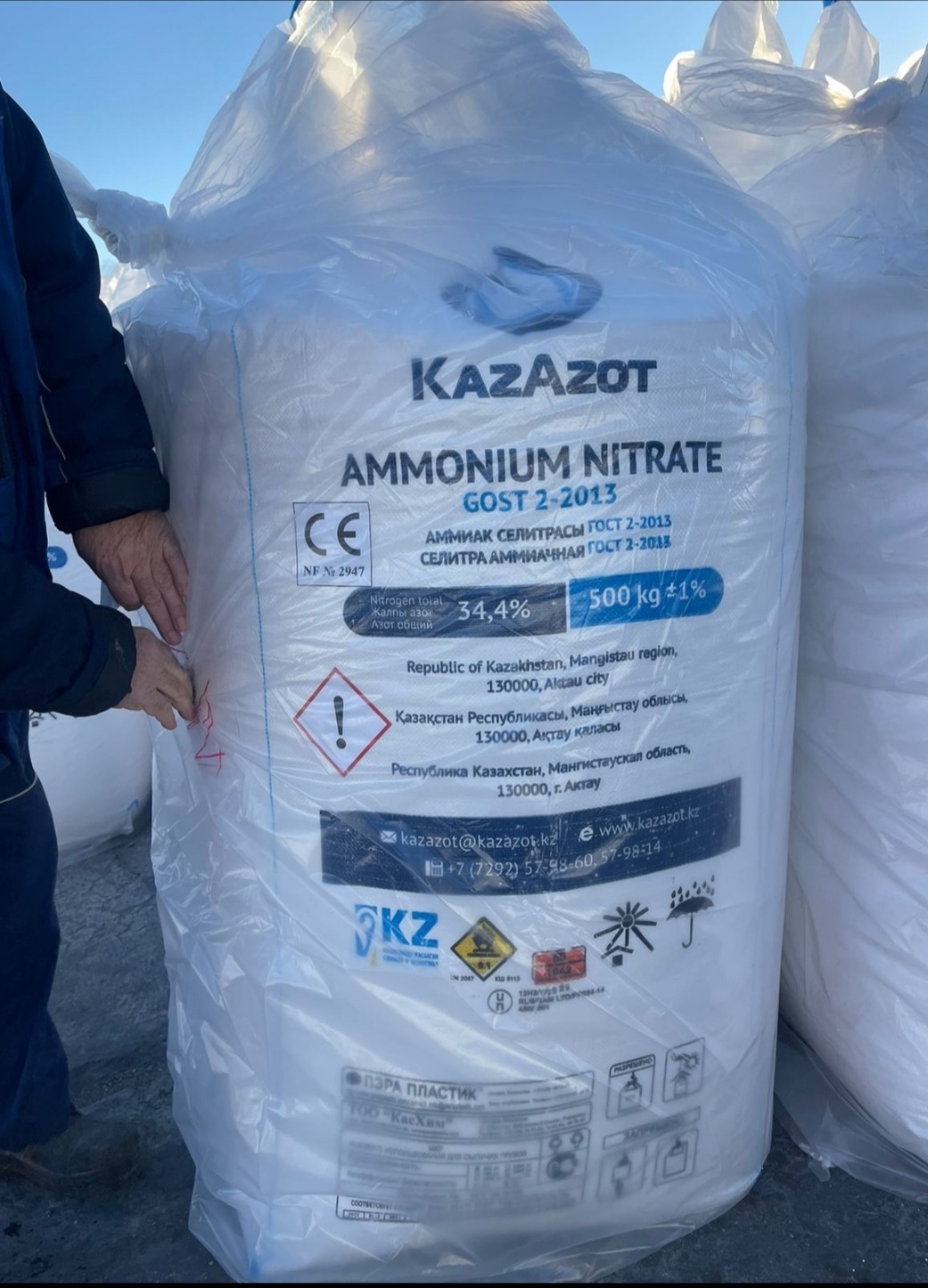 Duży, biały, plastikowy worek z granulowanym azotem amonowym KAZAZOT 34.4%, zapakowany w Kazachstanie, z widocznymi oznaczeniami i certyfikatami, trzymany przez osobę w roboczym ubraniu.