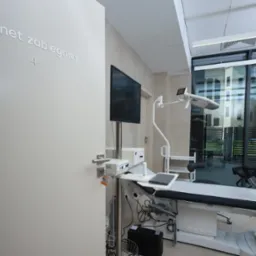 Wnętrze gabinetu zabiegowego z nowoczesnym kozetką ginekologiczną firmy Golem, lampą zabiegową, monitorem na statywie i dużym oknem z widokiem na zewnątrz.