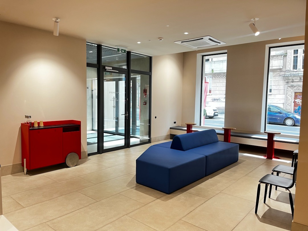 Jasne wnętrze poczekalni z niebieską sofą modułową, czerwonym wózkiem na kółkach z napojami i widokiem na ulicę przez duże okna.