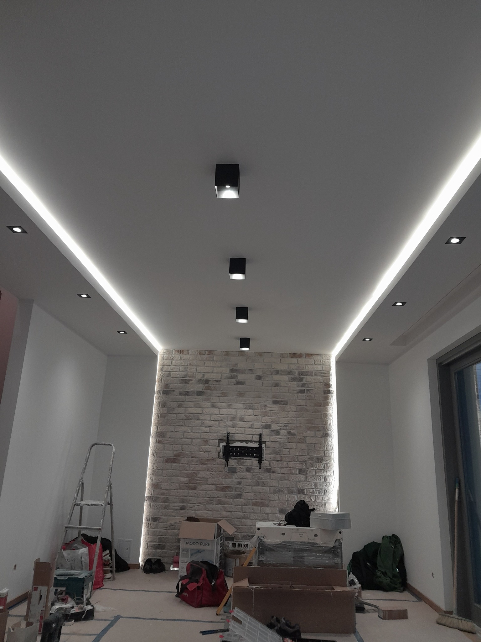 Nowoczesne oświetlenie sufitowe z ledowymi listwami i punktowymi lampami w pomieszczeniu z ceglaną ścianą, widoczne narzędzia i materiały budowlane.