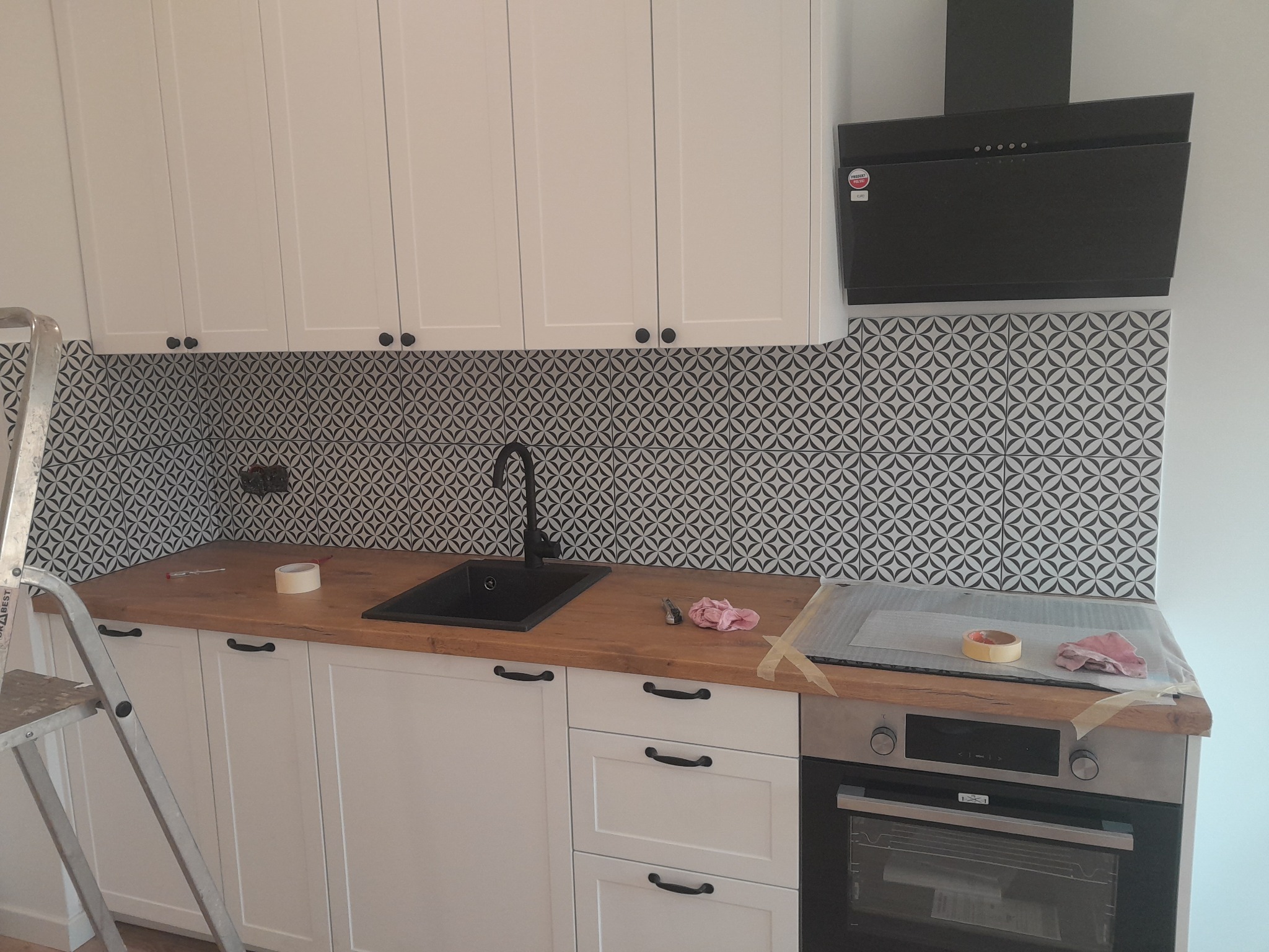 Kuchnia po remoncie: białe szafki z czarnymi uchwytami, drewniany blat, czarny zlew i bateria, wzorzyste płytki na ścianie, czarny okap, drabina malarska po lewej.