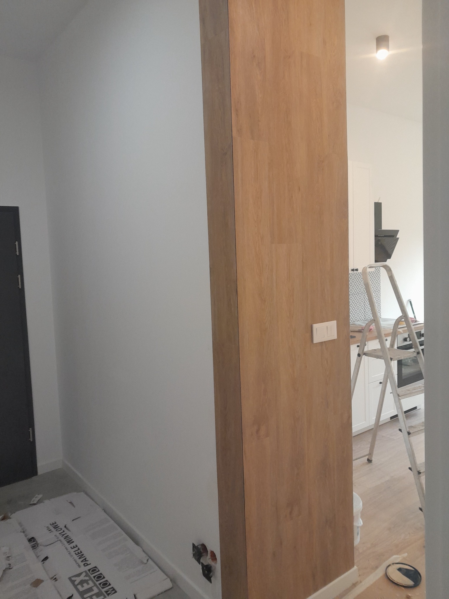 Wnętrze w trakcie remontu: świeżo pomalowana biała ściana, drewniana kolumna z kontaktem, kartony paneli podłogowych przy ścianie, widoczne gniazdka elektryczne, drabina malarska w tle.