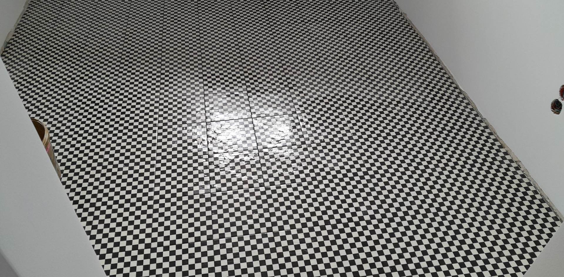 Ukończona podłoga w szachownicę z czarno-białych płytek ceramicznych, widok z góry, ściany pomalowane na biało, widoczne niedociągnięcia przy krawędziach ścian.