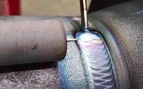 Detal spawu na metalowej rurze, widoczne tęczowe przebarwienia i elektroda w trakcie pracy.