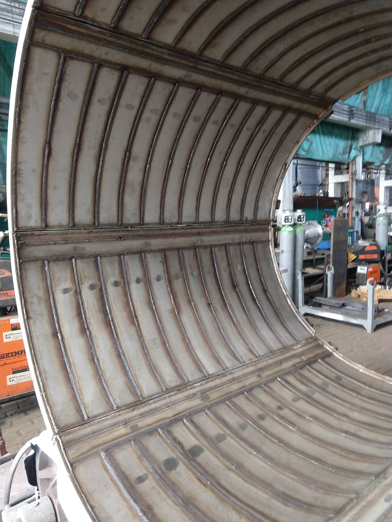 Metalowa konstrukcja łukowa ze śladami spawania, widoczne spoiny i elementy wzmacniające, w tle warsztat z butlami gazowymi i sprzętem spawalniczym.