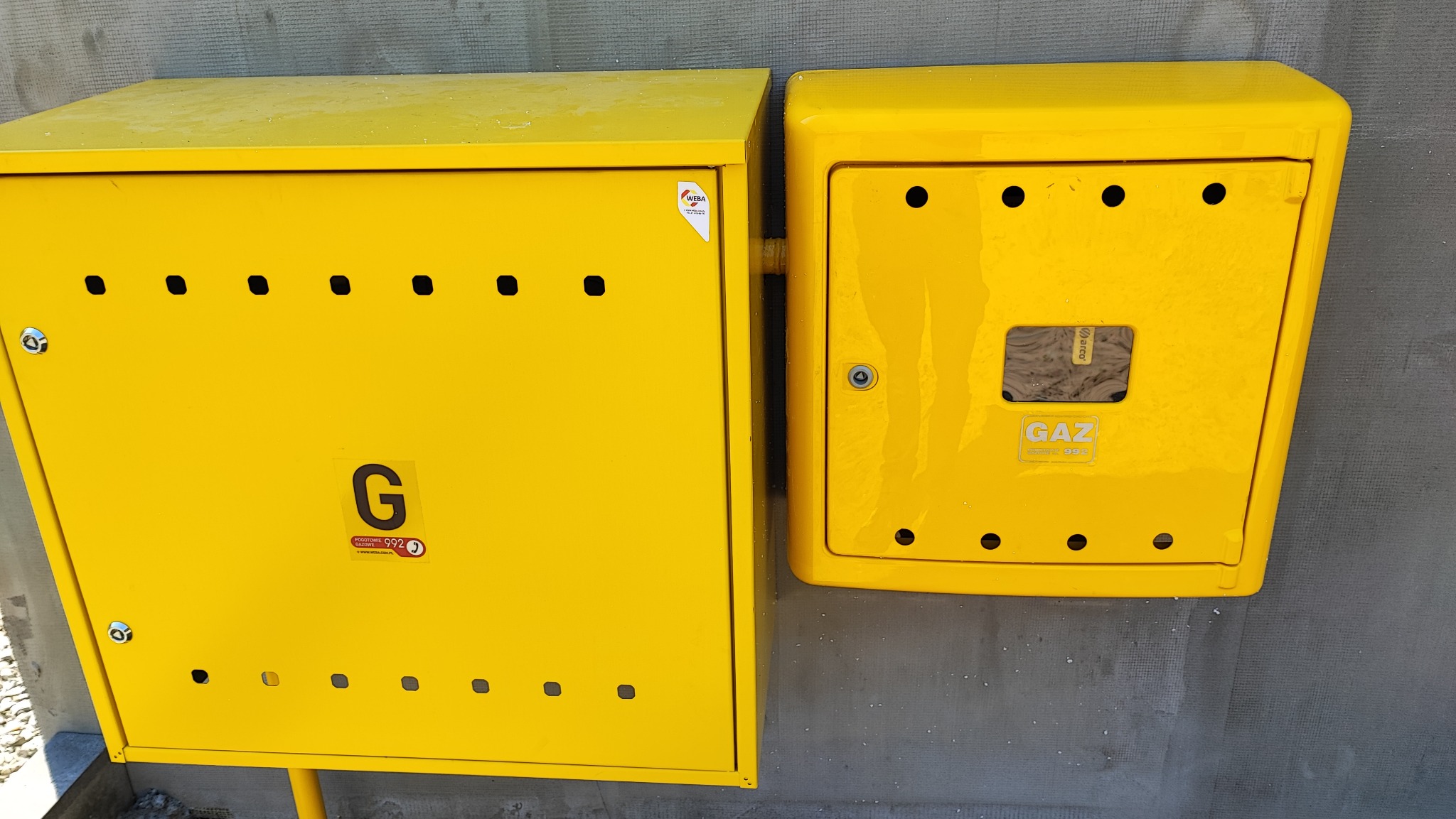 Dwie żółte skrzynki gazowe zamontowane na szarej ścianie, większa z otworem wentylacyjnym i oznaczeniem 'G', mniejsza z zamkiem i napisem 'GAZ'.