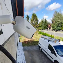 Zamontowana kamera monitoringu Dahua na elewacji budynku, widoczny przewód, w tle biały bus z drabiną na dachu i fragment ulicy. Błękitne niebo z chmurami.