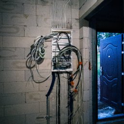 R-TECH Dawid Rogowski - Rozdzielnia elektryczna z licznymi przewodami w surowym wnętrzu. Widoczne kable prowadzone po ścianie z bloczków, otwarte drzwi wejściowe. Instalacja w trakcie prac.