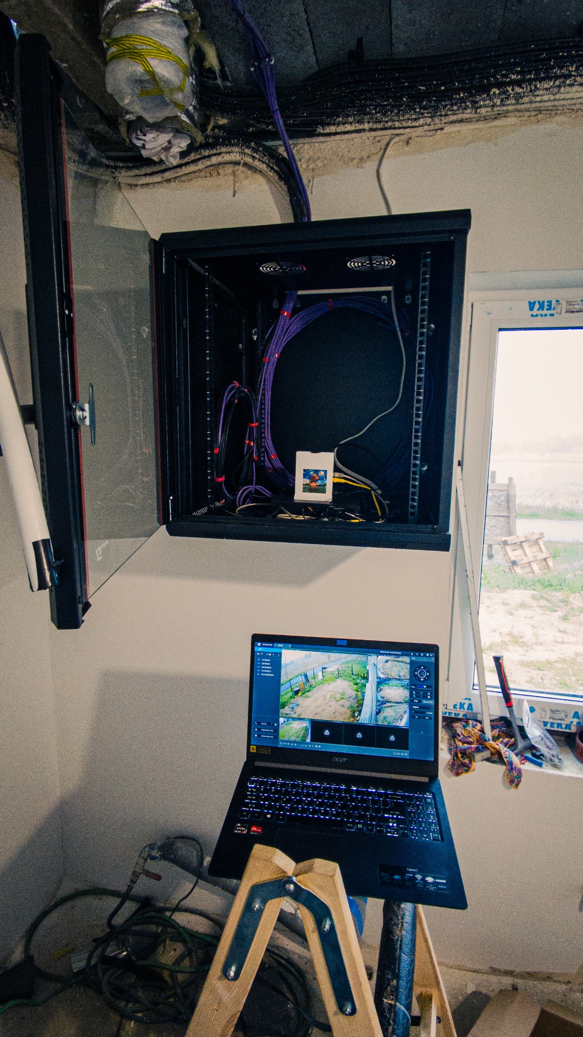 Uruchomiony system monitoringu na budowie: laptop z podglądem z kamer, szafa rack z okablowaniem. Widok z okna na zewnątrz.