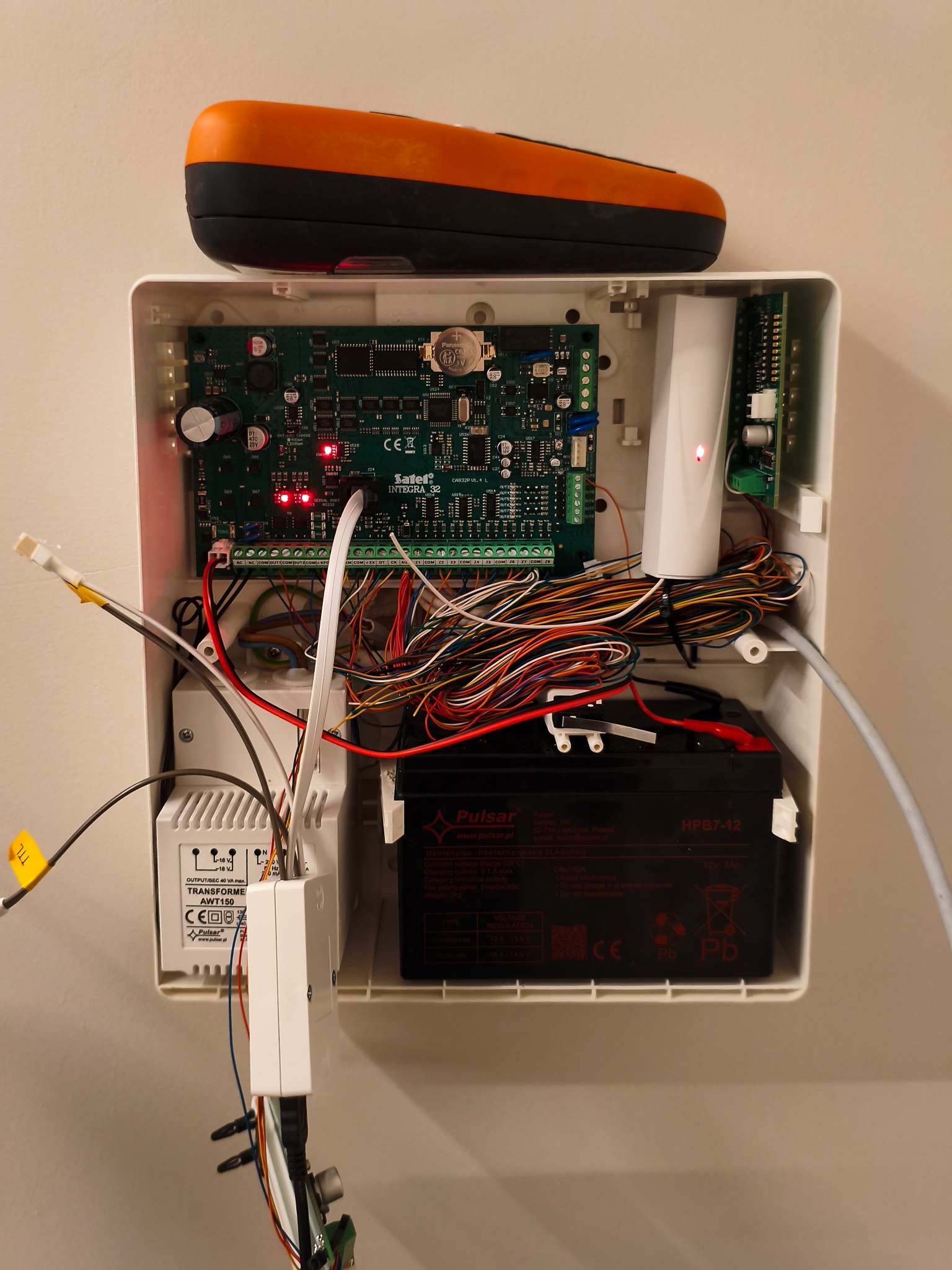 Otwarta obudowa centrali alarmowej Satel Integra 32 z widoczną elektroniką, akumulatorem Pulsar HPB7-12 i transformatorem AWT150. Na górze leży pomarańczowy miernik.