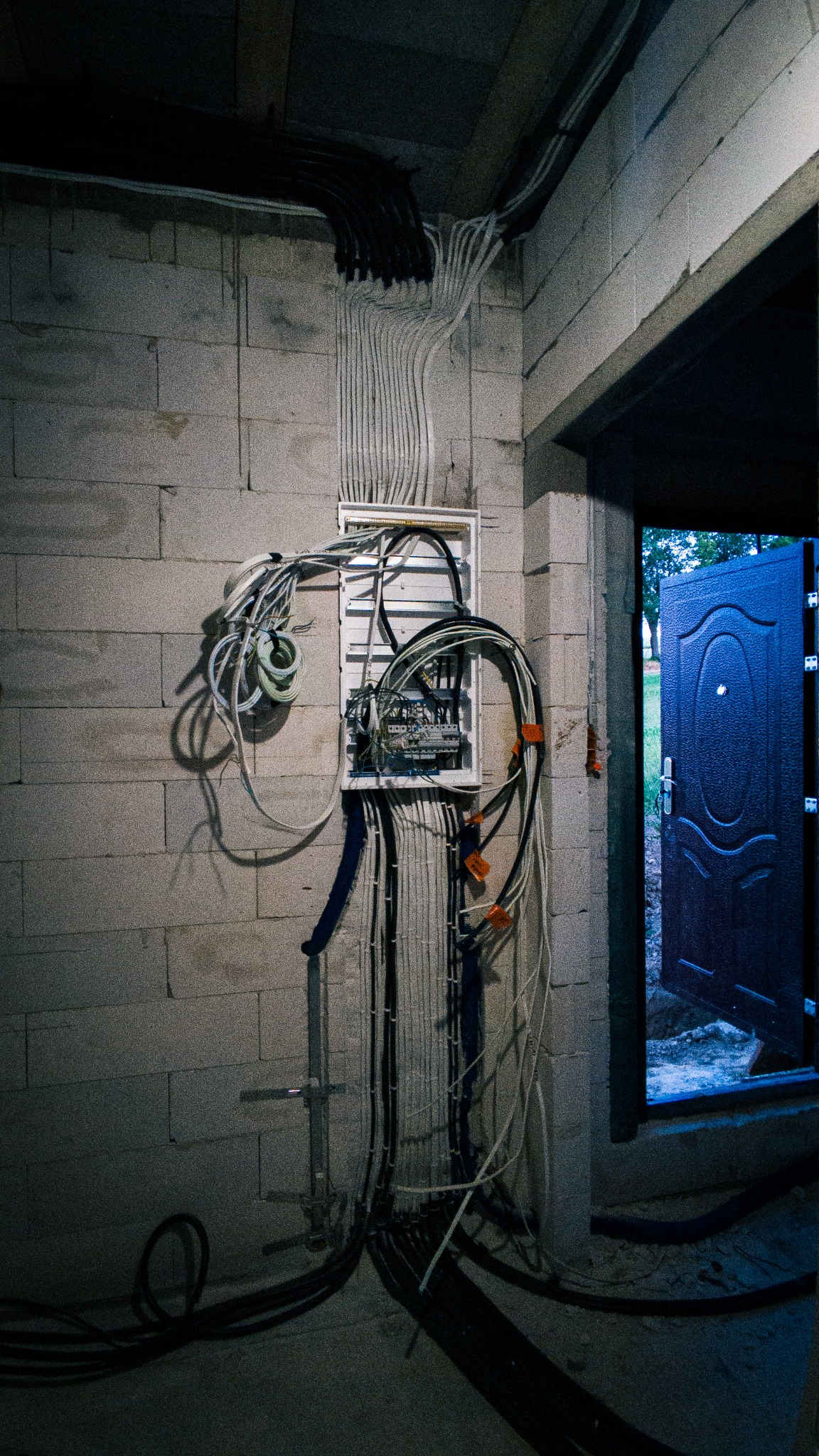 Rozdzielnia elektryczna z licznymi przewodami w surowym wnętrzu. Widoczne kable prowadzone po ścianie z bloczków, otwarte drzwi wejściowe. Instalacja w trakcie prac.