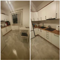 Uporządkowana kuchnia z białymi szafkami z czarnymi uchwytami i drewnianym blatem, widok na stół z krzesłami i okno z firanką. Na blacie czajnik, płyta indukcyjna i frytkownica beztłuszczowa.