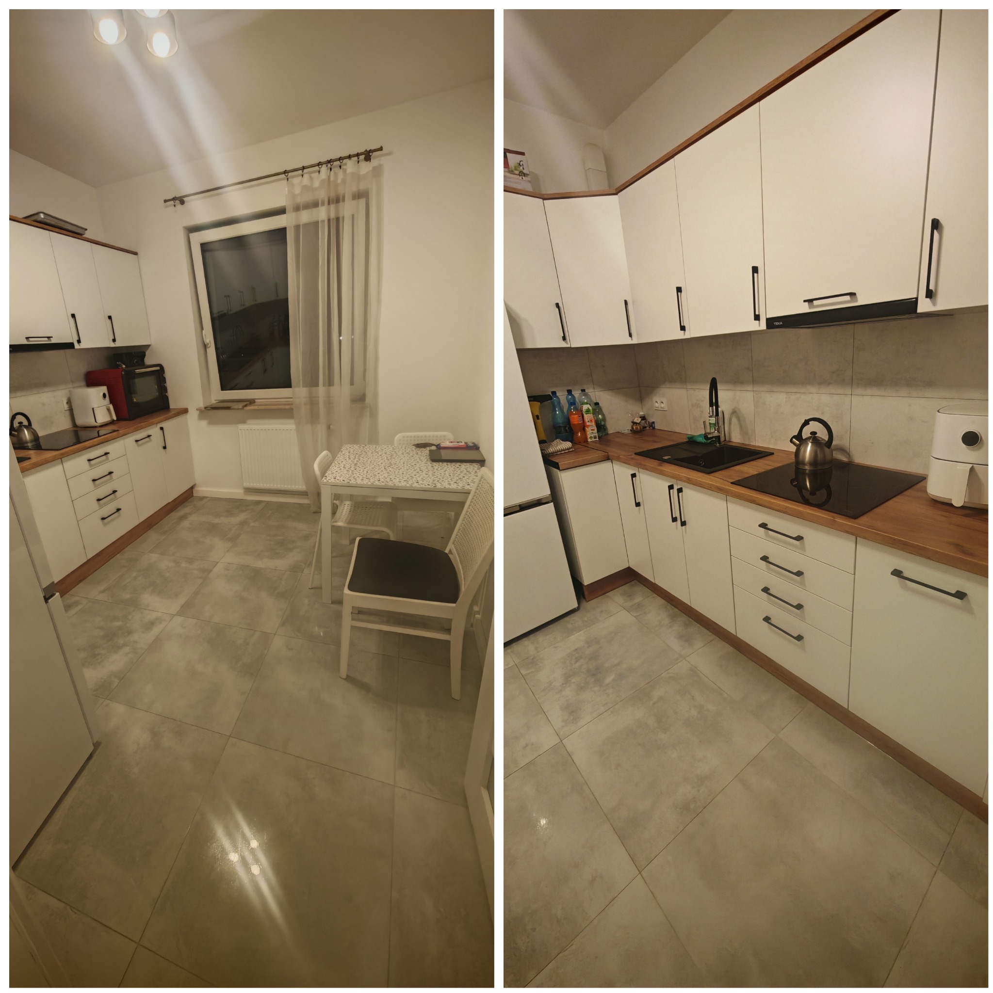 Uporządkowana kuchnia z białymi szafkami z czarnymi uchwytami i drewnianym blatem, widok na stół z krzesłami i okno z firanką. Na blacie czajnik, płyta indukcyjna i frytkownica beztłuszczowa.