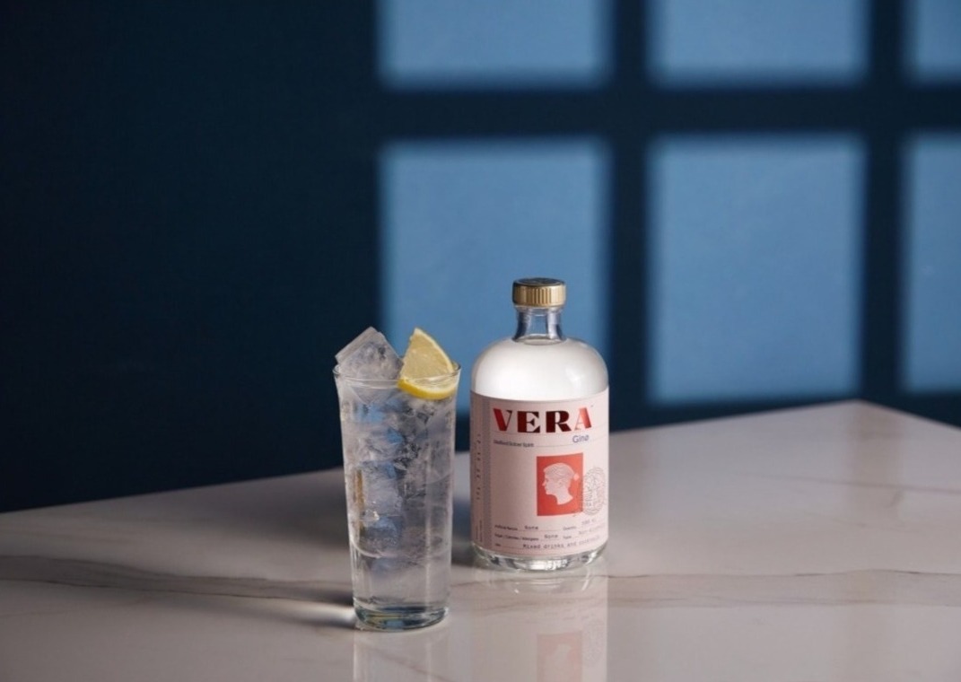 Bezalkoholowy gin Vera Gino w butelce obok szklanki z lodem i cytryną, na marmurowym blacie, z cieniem okna na ścianie w tle.