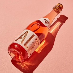 Butelka bezalkoholowego, różowego wina musującego ALT Drinks na jasnoróżowym tle, rzucająca wyraźny cień.