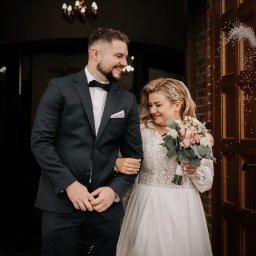 PATRYCJA SZMIDT PAAPITA FOTOGRAFIA - Uśmiechnięta para młoda wychodzi z kościoła, obsypywana ryżem. Panna młoda trzyma bukiet, a pan młody ma muchę. Widoczny fragment ceglanej ściany i drzwi.