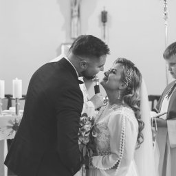 PATRYCJA SZMIDT PAAPITA FOTOGRAFIA - Czarno-białe ujęcie pary młodej podczas ceremonii ślubnej w kościele, z ministrantem w tle. Panna młoda trzyma bukiet, a pan młody mówi do mikrofonu.