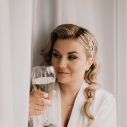 PATRYCJA SZMIDT PAAPITA FOTOGRAFIA - Panna młoda w białym szlafroku, z upiętymi blond włosami i delikatną biżuterią, trzyma kieliszek białego wina, spoglądając w bok przy oknie z firanką.