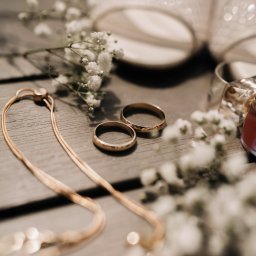 PATRYCJA SZMIDT PAAPITA FOTOGRAFIA - Dwa złote obrączki ślubne na drewnianym tle, otoczone drobnymi białymi kwiatami i elementami biżuterii, z delikatnym oświetleniem i butami ślubnymi w tle.
