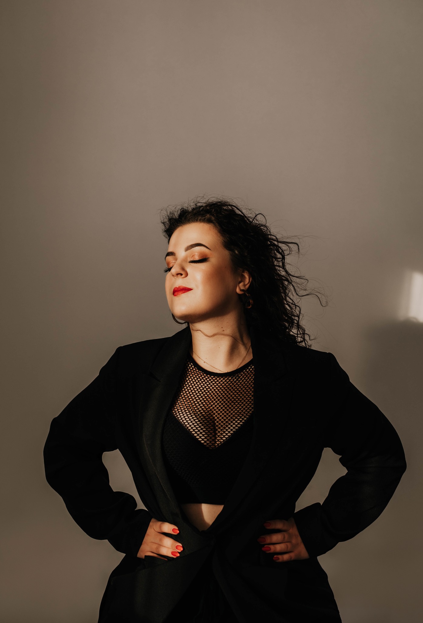 Portret kobiety w czarnym żakiecie, siateczkowej bluzce i czerwonej szmince, pozującej z zamkniętymi oczami na neutralnym tle, oświetlonej bocznym światłem.