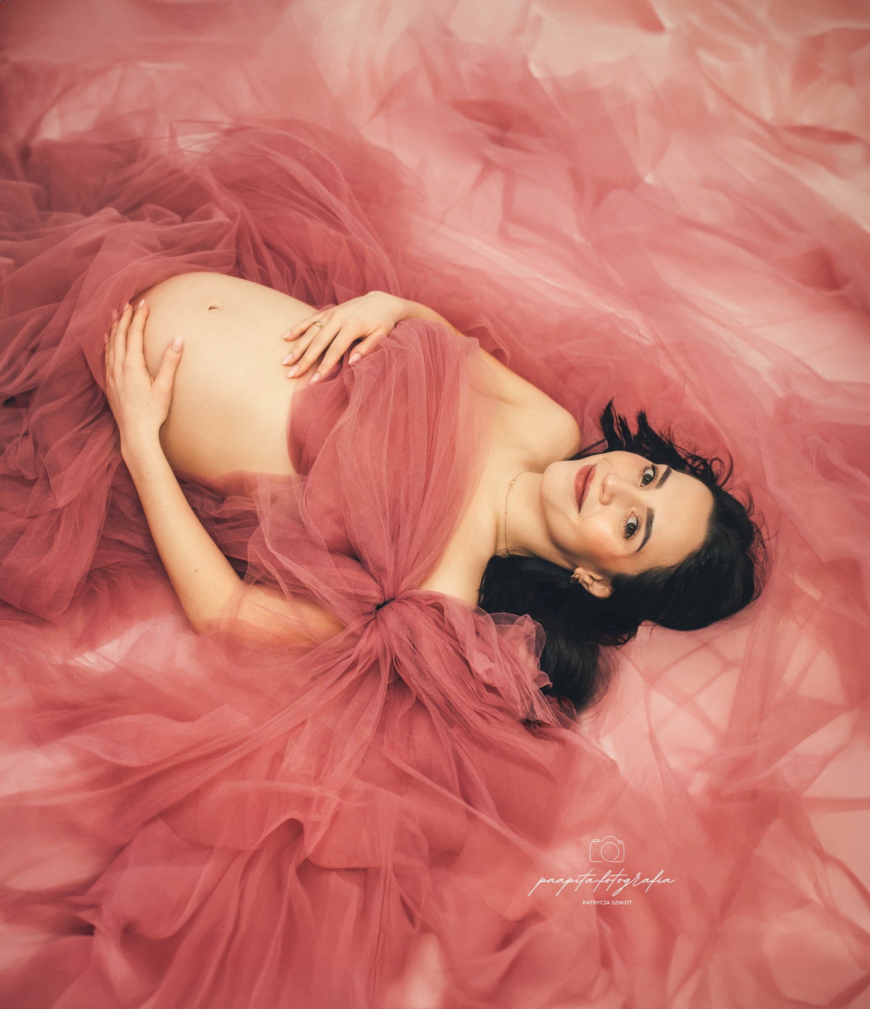 Kobieta w ciąży, leżąca na różowym tiulu, z rękami na brzuchu, uśmiechnięta, z widocznym pierścionkiem na palcu.