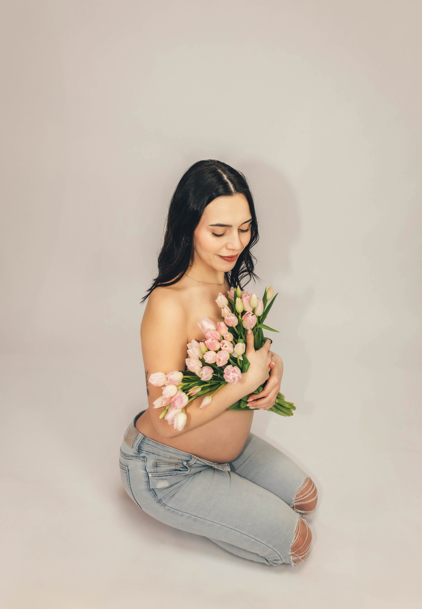 Kobieta w ciąży, siedząca na kolanach, zakrywająca piersi bukietem różowych tulipanów, ubrana w podarte jasne dżinsy, na jasnym tle.