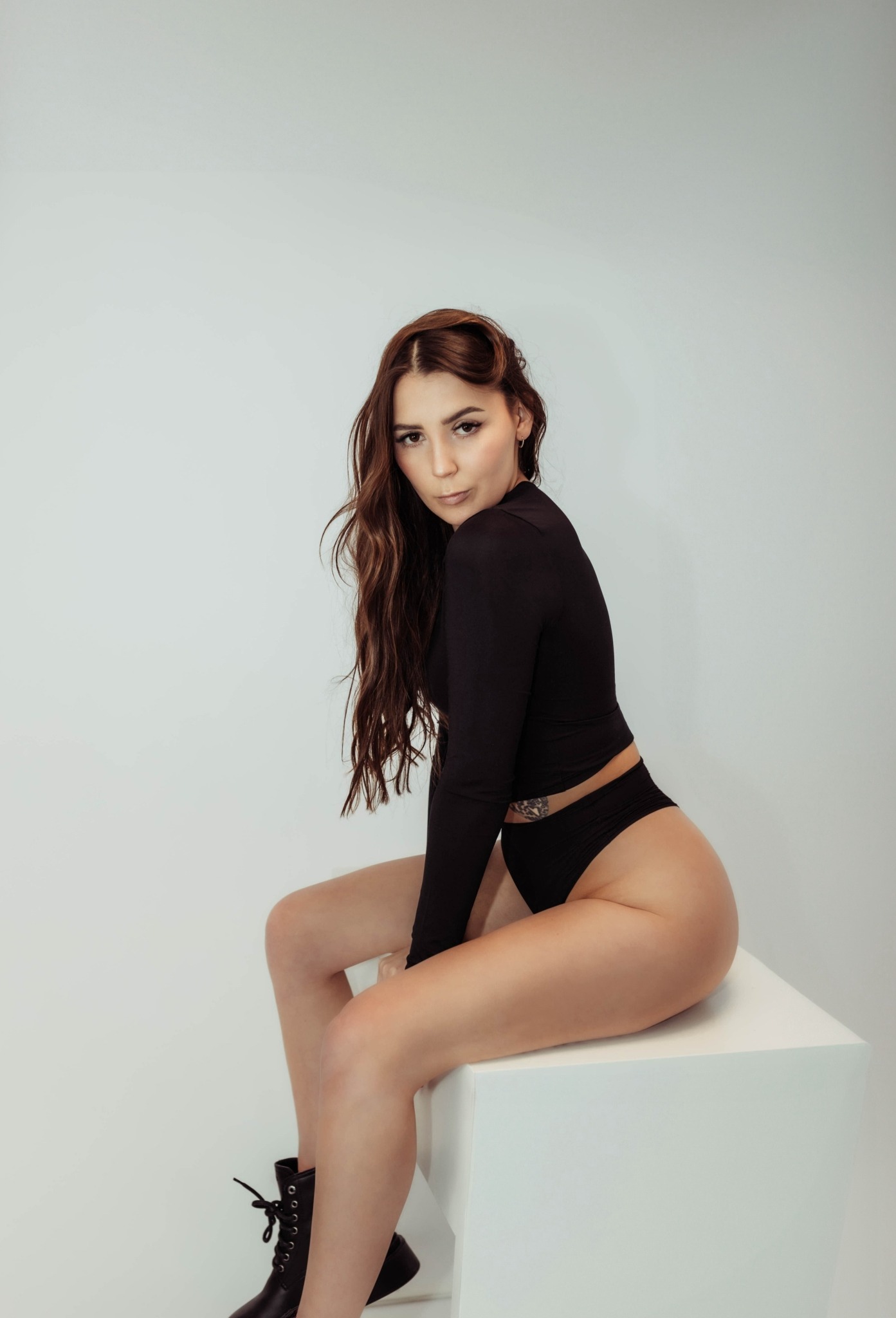 Brunetka w czarnym body i botkach siedzi na białym sześcianie w studiu, spoglądając przez ramię. Widoczne jest tatuaż na boku i delikatny makijaż.