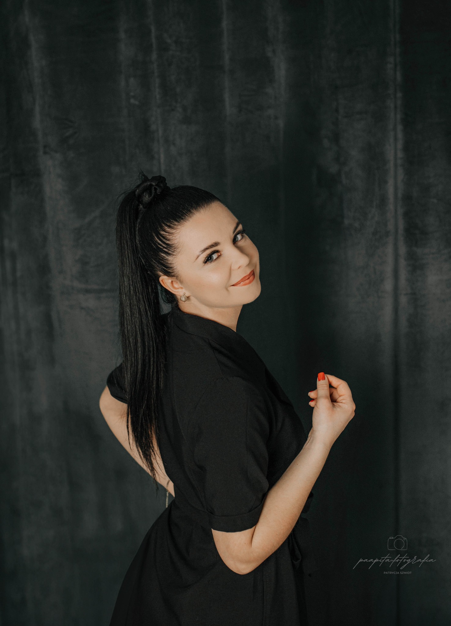 Portret kobiety w czarnej sukience, odwróconej przez ramię, z włosami upiętymi w wysoki kucyk na tle ciemnego, teksturowanego tła.