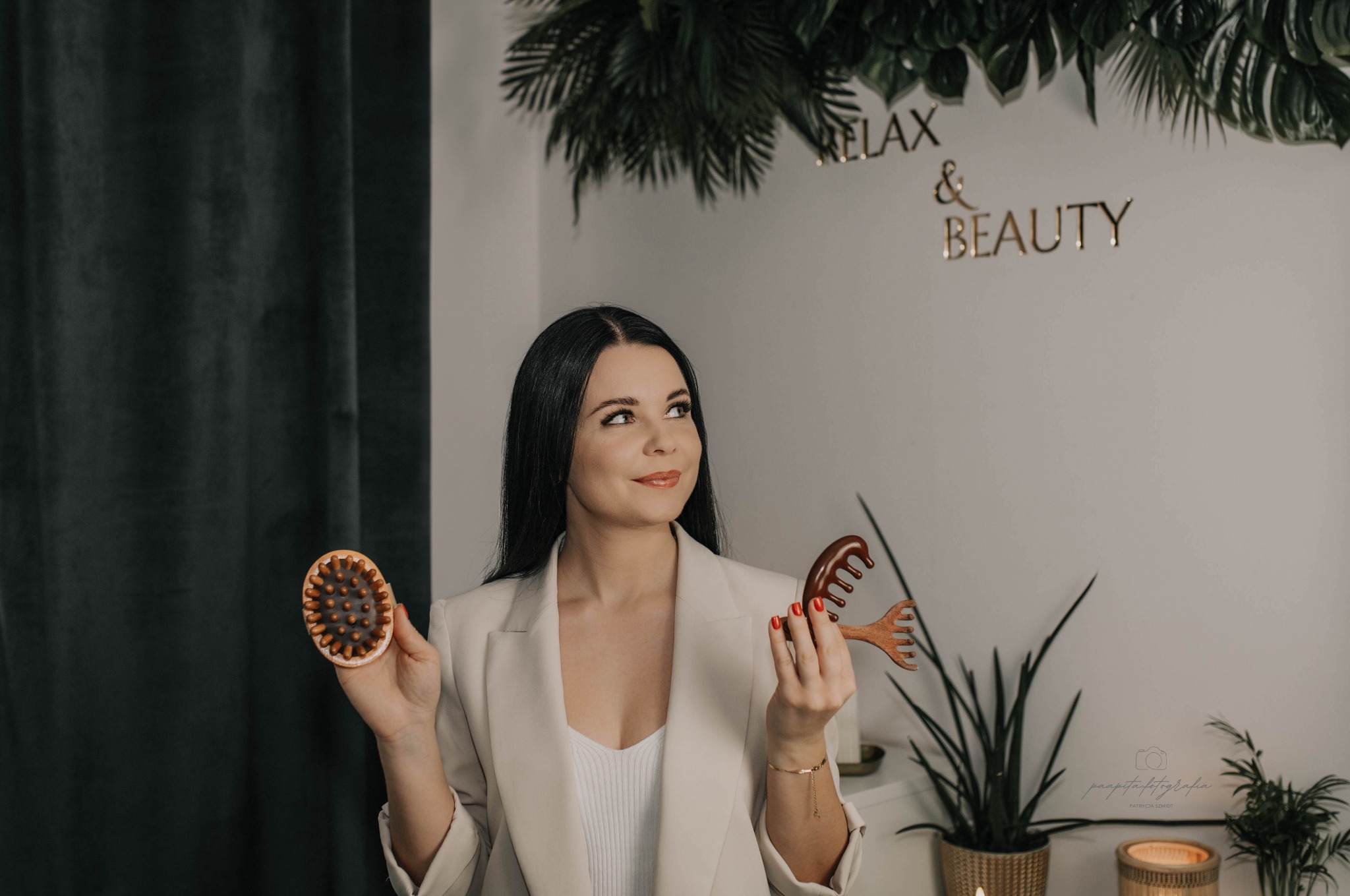 Brunetka w beżowej marynarce prezentuje drewnianą szczotkę i grzebień na tle ściany z napisem 'Relax & Beauty' i roślinami