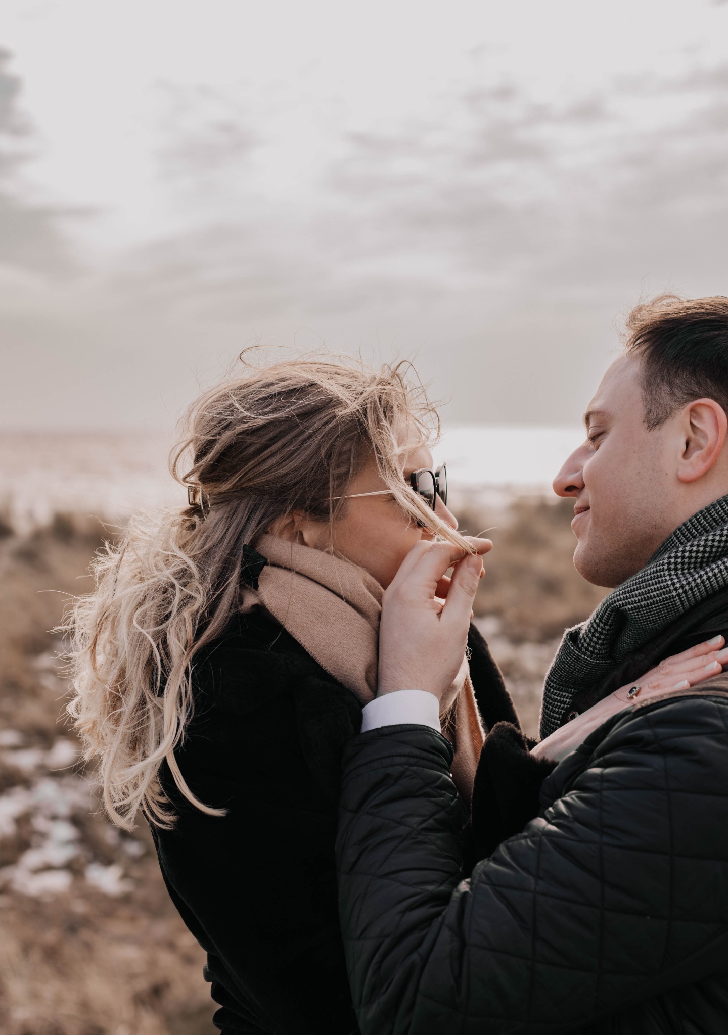 Zakochana para na tle plaży; mężczyzna delikatnie bawi się kosmykiem włosów kobiety w okularach przeciwsłonecznych.
