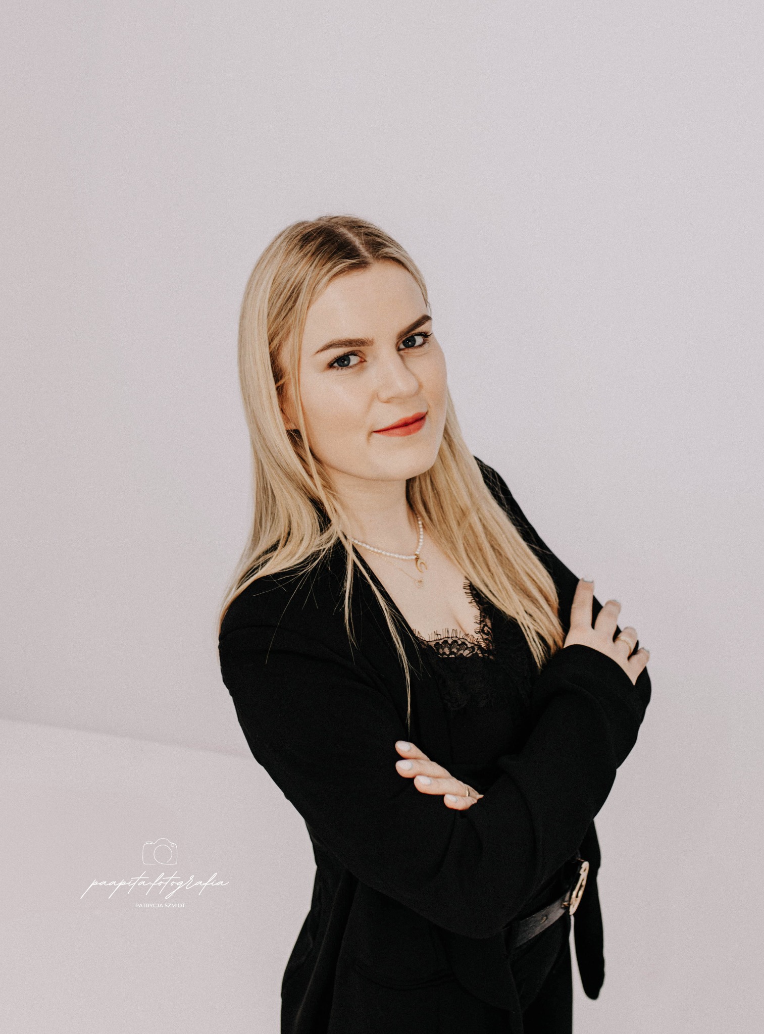 Portret młodej kobiety z blond włosami, ubranej w czarny żakiet i koronkową bluzkę, pozującej na jasnym tle z założonymi rękami.