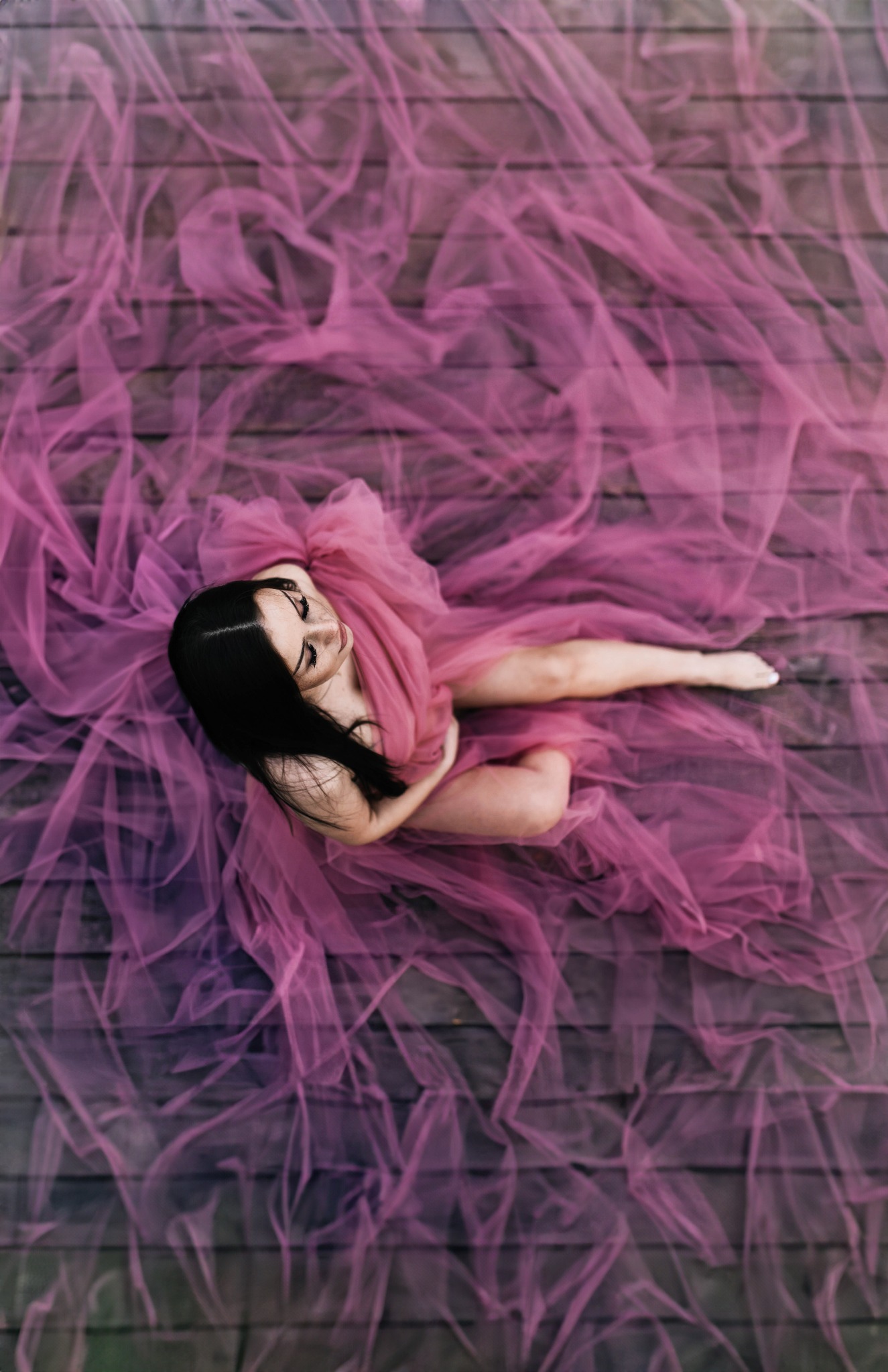 Kobieta leżąca na drewnianej podłodze, otulona różowym, transparentnym materiałem, widok z góry.