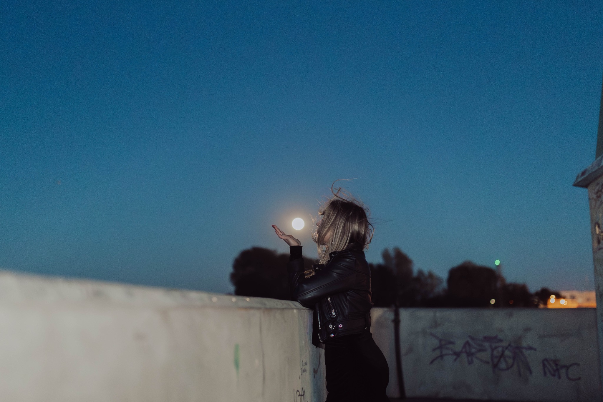 Kobieta w skórzanej kurtce, stojąc na tle nocnego nieba, dmucha w dłonie w kierunku księżyca, tworząc iluzję trzymania go.